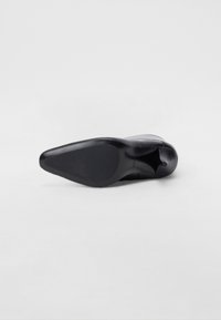 Chaussure noire à talons hauts vue de dessous. Présente un embout pointu et une finition lisse et brillante avec une semelle légèrement texturée.