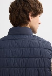 Chaleco acolchado azul marino con líneas horizontales de acolchado y un cuello alzado. Textura suave, material ligero, adecuado para superponer.