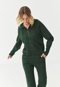 Sudadera verde con cremallera en tejido suave, con un bolsillo frontal, pantalones a juego y capucha. Presenta un corte holgado y puños de canalé.