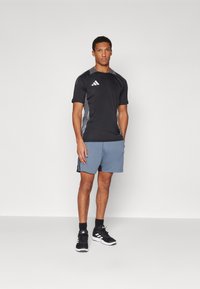Sportbekleidung mit einem schwarzen kurzärmeligen Shirt mit grauen Seiten und blauen Shorts. Schwarze Sneakers runden den Look ab.