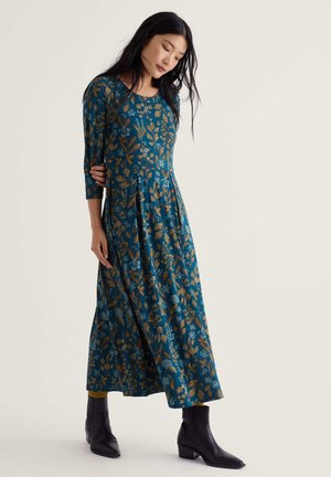 Femme portant une longue robe bleu sarcelle avec un motif floral marron et bleu, associée à des bottines noires, debout devant un fond uni.