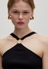 Zwarte geribbelde haltertop met een gouden hardwaredetail bij de halslijn. Model draagt grote gouden oorbellen. Neutrale achtergrond.