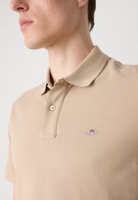 Polo beige en tissu texturé avec un col rabattu, deux boutons et un petit logo brodé sur la poitrine.