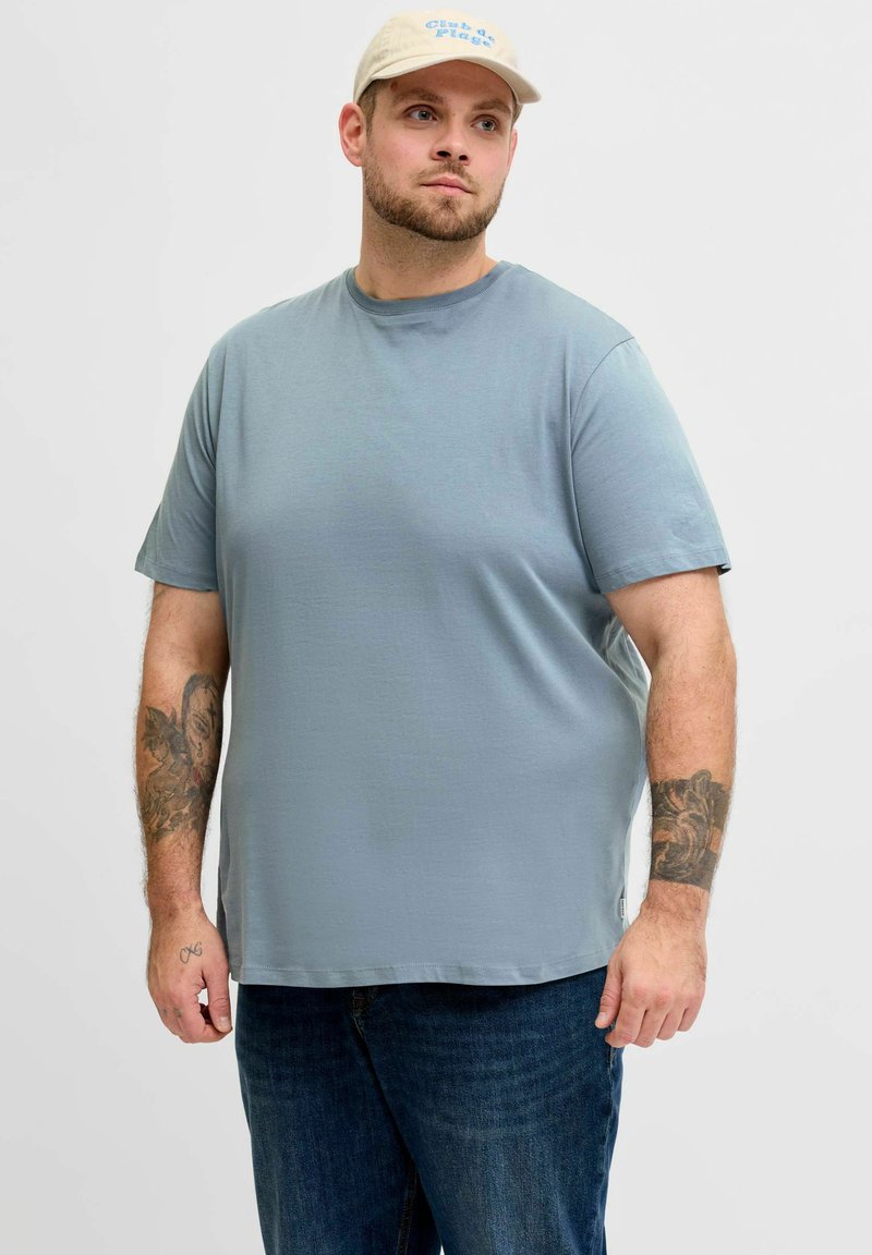 Mees helesinises T-särgis ja tumedates teksapükstes kannab beežikat nokamütsi, näidates mõlema käsivarre tätoveeringuid, seisab ühevärvilise heleda tausta ees.