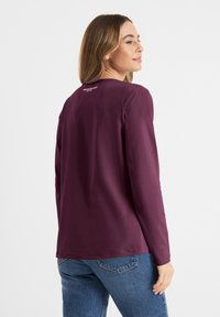 Langarm t-shirt in diep paars, gemaakt van zachte stof. Heeft een ronde halslijn en een klein logo op de achterkant. Gladde textuur.