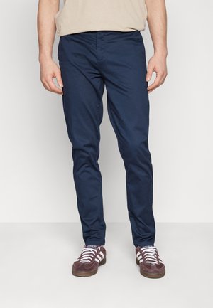 Mężczyzna ubrany w granatowe, dopasowane spodnie i brązowe buty sportowe Adidas z białymi paskami, stojący na jednolitym białym tle.