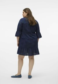 Navyblaues Kleid mit Lochmuster, 3/4-Ärmeln und rüschengem Saum. Kombiniert mit blauen Ballerinas, wird das Modell von hinten gezeigt.