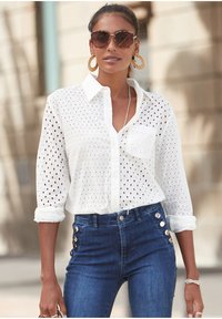 Camicia bianca con ritagli ad occhio, maniche lunghe arrotolate, abbinata a jeans blu a vita alta con dettagli in bottone dorato.