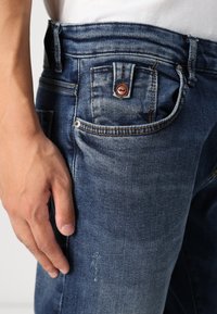 Vaqueros de denim en azul oscuro con un acabado desgastado, que presentan costuras visibles y un botón marrón en el bolsillo frontal. Tejido texturizado.