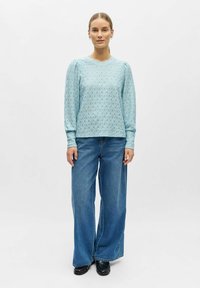 Lys blå langermet topp med et teksturert blomstermønster, kombinert med vide blå denimjeans. Modellen står foran en nøytral bakgrunn.