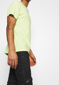 Man som bär en ljusgrön kortärmad skjorta, svarta byxor med dragkedjedetalj, silvrigt kedjesmycke och en arm-tatuering med romerska siffror.