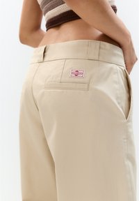 Pantalons en coton beige avec une coupe droite, dotés d'une poche arrière et d'une étiquette rouge et blanche près de la taille. Texture lisse.