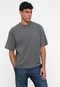 Grå kortermet T-skjorte laget av bomull med rund hals. Har en liten logo i lys grønn på brystet. Kombinert med jeans.