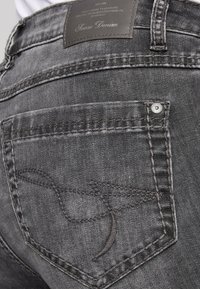 Pantalones vaqueros de mezclilla negra con un bolsillo trasero con costuras curvas y una etiqueta de cuero que muestra el nombre de la marca, con un acabado desgastado.