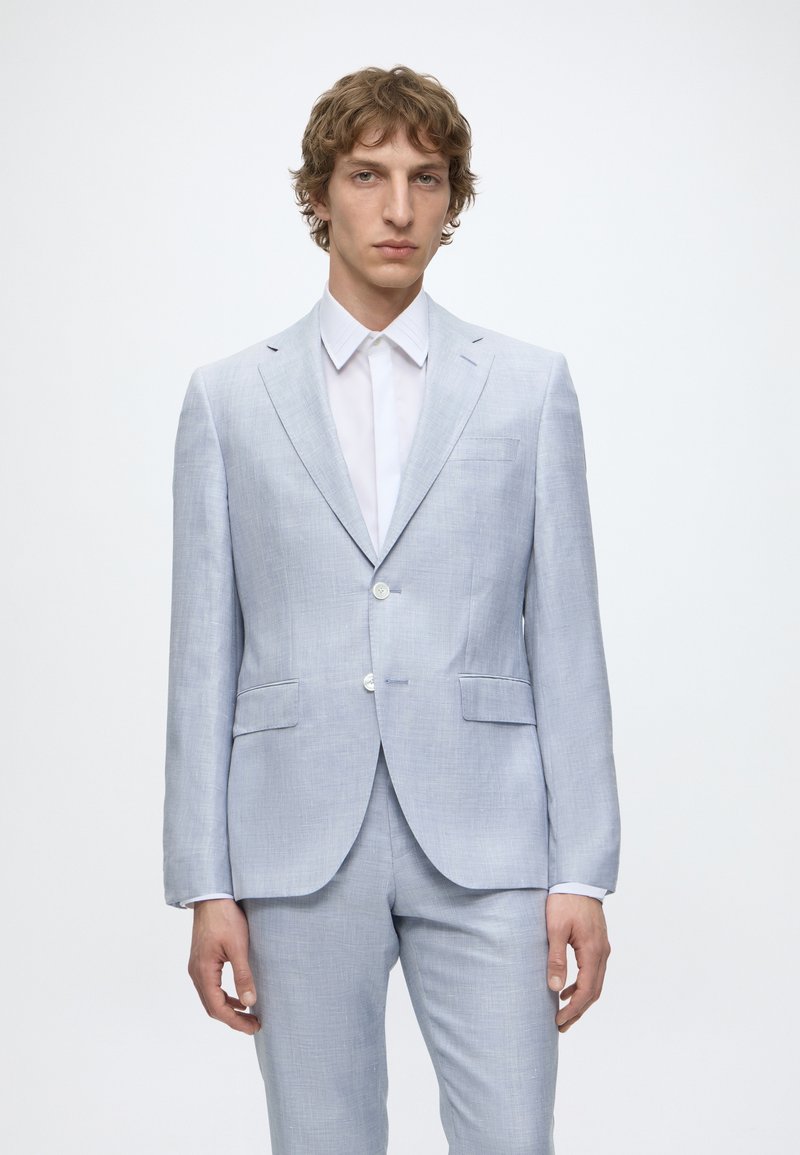Jeune homme portant un costume gris clair avec des boutons blancs et une chemise blanche, debout devant un fond blanc uni.