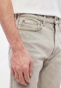 Gaiši bēšas džinsa bikses ar nelielu nēsātu efektu, uz priekšējā kabatas atzīmes ar sarkanu un baltu Calvin Klein logo un metāla kniedēm.