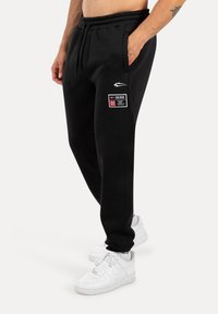 Zwarte sweatpants met een elastische tailleband, zijzakken en een patch met witte tekst. Gecombineerd met witte sportschoenen.