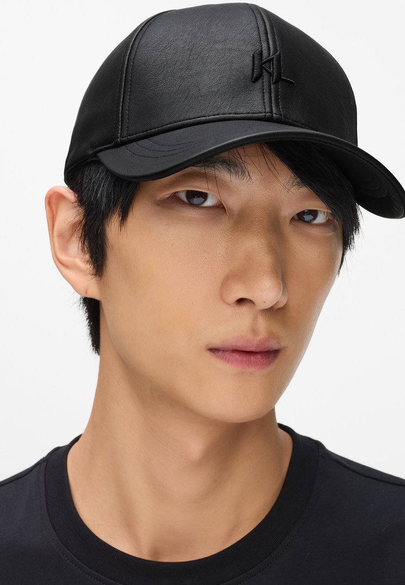Černá kožená čepice s prohnutým kšiltem a prošívanými detaily. Na přední straně je decentní vyšívané logo. Hladký povrch.