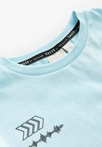 Šviesiai mėlynas medvilnės marškinėlis su apvaliu iškirpimu, juoda etiketė su užrašu "BAKER by TED BAKER" ir grafiniu dizainu, kuriame pavaizduoti rodyklės ir garso bangos.