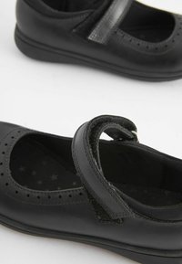 Zapatos de cuero negros con punta redondeada, diseño perforado y una correa ajustable. Forro interior texturizado con patrón de estrellas. Suela de goma.