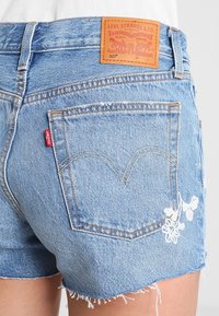 Pantalones cortos de mezclilla Levi's de color azul claro con un bolsillo trasero, etiqueta roja, parche de cuero de la marca y bordado floral blanco cerca del dobladillo.
