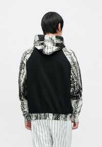 Bluza z kapturem o czarnym korpusie i abstrakcyjnym wzorze na rękawach w odcieniach bieli i szarości. Charakteryzuje się luźnym krojem i ściągaczami na dole i przy mankietach.