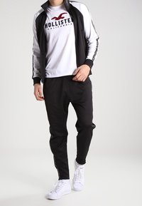 Svart träningsjacka med vita detaljer över en vit T-shirt med "Hollister"-logotyp. Svarta joggerbyxor och vita sneakers.