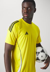 T-shirt de sport jaune avec des rayures noires sur les épaules, coupe ajustée, et logo sur la poitrine. Un ballon de soccer noir et blanc est tenu sous un bras.