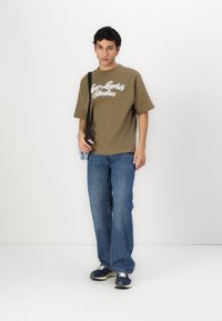 Oliven grøn oversized t-shirt med hvid tekst "Low Lights Studios", parret med lyseblå jeans og marineblå sneakers, bærende en lille blå taske.