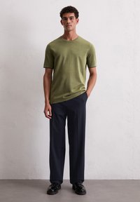 Zielona oliwkowa koszulka z krótkim rękawem i okrągłym dekoltem, w połączeniu z szerokimi granatowymi spodniami i czarnymi butami, na neutralnym tle.