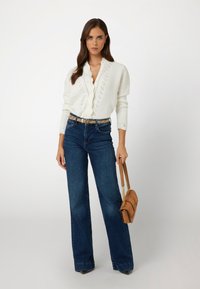Cardigan bianco lavorato a maglia con bordo ondulato, abbinato a jeans flare in denim blu a vita alta e una borsa strutturata marrone.