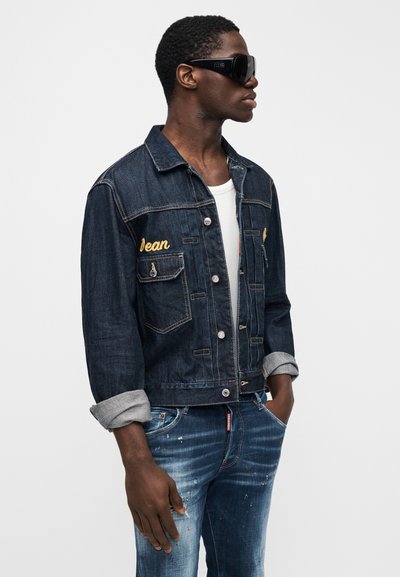 Uomo con giacca di jeans scura con ricamo "Jean", maniche arrotolate, camicia bianca, jeans blu consumati e grandi occhiali da sole neri, in piedi di profilo.