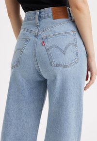 Jean en denim Levi's bleu clair taille haute avec poches arrière, patch en cuir de la marque et étiquette rouge Levi's visible sur la poche droite, porté par une personne.