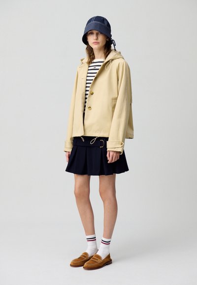Veste beige imperméable avec capuche et deux boutons, portée par-dessus une chemise rayée bleu marine, associée à une jupe plissée bleu marine et des mocassins marron.