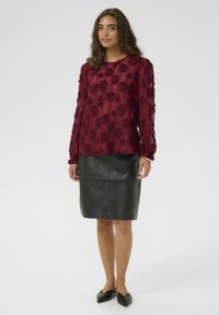 Blusa trasparente color borgogna con motivo floreale testurizzato, maniche lunghe e polsini arricciati, abbinata a una gonna in pelle nera aderente.