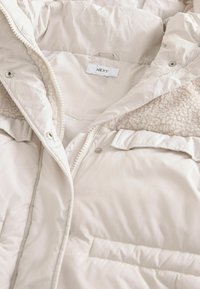 Dik beige jack met een zachte fleece voering, sluiting met knopen en rits, en een label met de tekst "NEXT" aan de binnenkant. Het heeft een zak aan de voorkant.