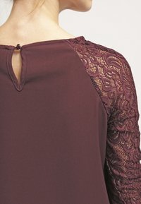 Burgundy paita, jossa on pitsihihat ja avara selkäosa. Pehmeä tekstuuri, jossa on monimutkainen pitsikuvio hartioilla.