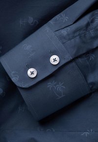 Manga de camisa azul marino con sutiles patrones de palmeras y flamencos, botones plateados y costuras en contraste en el puño.