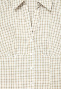 Chemise à carreaux beige et blanche, boutonnée, avec col, taille cintrée et manches courtes.