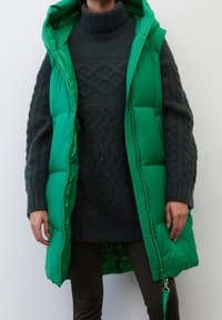 Grøn puffer vest med hætte, med en quiltet design og lynlås lukning. Båret over en mørk striktrøje med ribbedeærmer og kant.