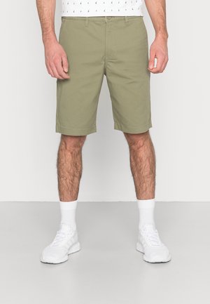 Homme portant un short vert olive clair jusqu'aux genoux, des chaussettes blanches mi-mollet et des chaussures de sport blanches, debout sur un sol gris.