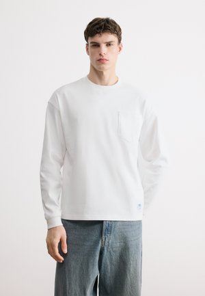 Mladý muž stojí čelem dopředu, má na sobě bílé tričko s dlouhými rukávy a náprsní kapsou a světle modré džínové kalhoty.