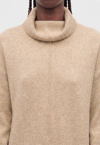 Person trägt einen beigen gestrickten Rollkragenpullover mit sichtbarer vertikaler Naht in der Mitte vorne und braunen geflochtenen Haaren.