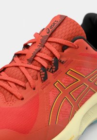 Red athletic shoe with a mesh upper, flat shoelaces, textured patterns, and contrasting yellow accents on the sides and sole.
