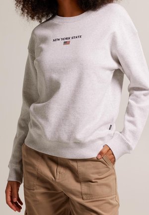 Person wearing light gray sweatshirt with "NEW YORK STATE" and a small American flag above beige pants, one hand in pocket.