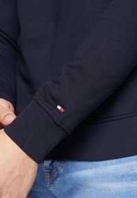 Námořnická mikina s žebrovanými manžetami, která má na levém zápěstí malou červenou, bílou a modrou značku. Hladký textilní povrch.