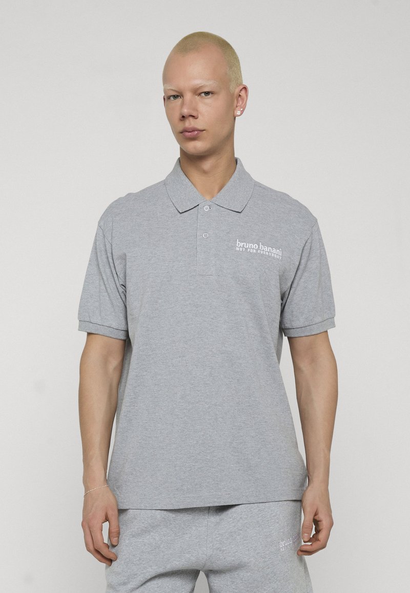 Personne aux cheveux blonds courts portant un polo gris et un pantalon gris assorti avec le texte "bruno banani" sur les deux.