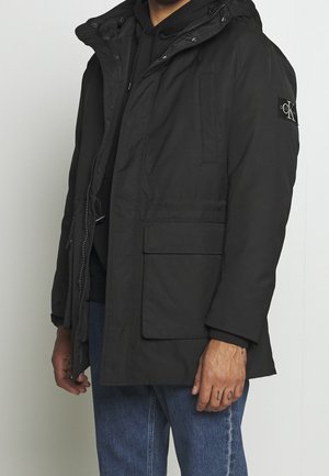 Black insulated parka jacket with a hood, zip front, two front pockets, and a logo patch on the sleeve; features ribbed cuffs and textured fabric.