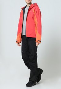 Veste imperméable rouge et orange avec pantalon noir, dotée d'une capuche, de fermetures éclair et de poches. Porté avec des chaussures de randonnée noires.