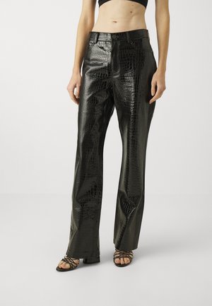 Mujer que lleva pantalones de piel sintética de cocodrilo negro brillante con botón y cremallera, combinados con sandalias de tacón negro con tiras.